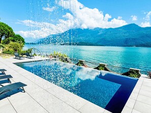 En swimmingpool med kristallklart vatten, omgiven av solstolar och med utsikt över en sjö och berg.