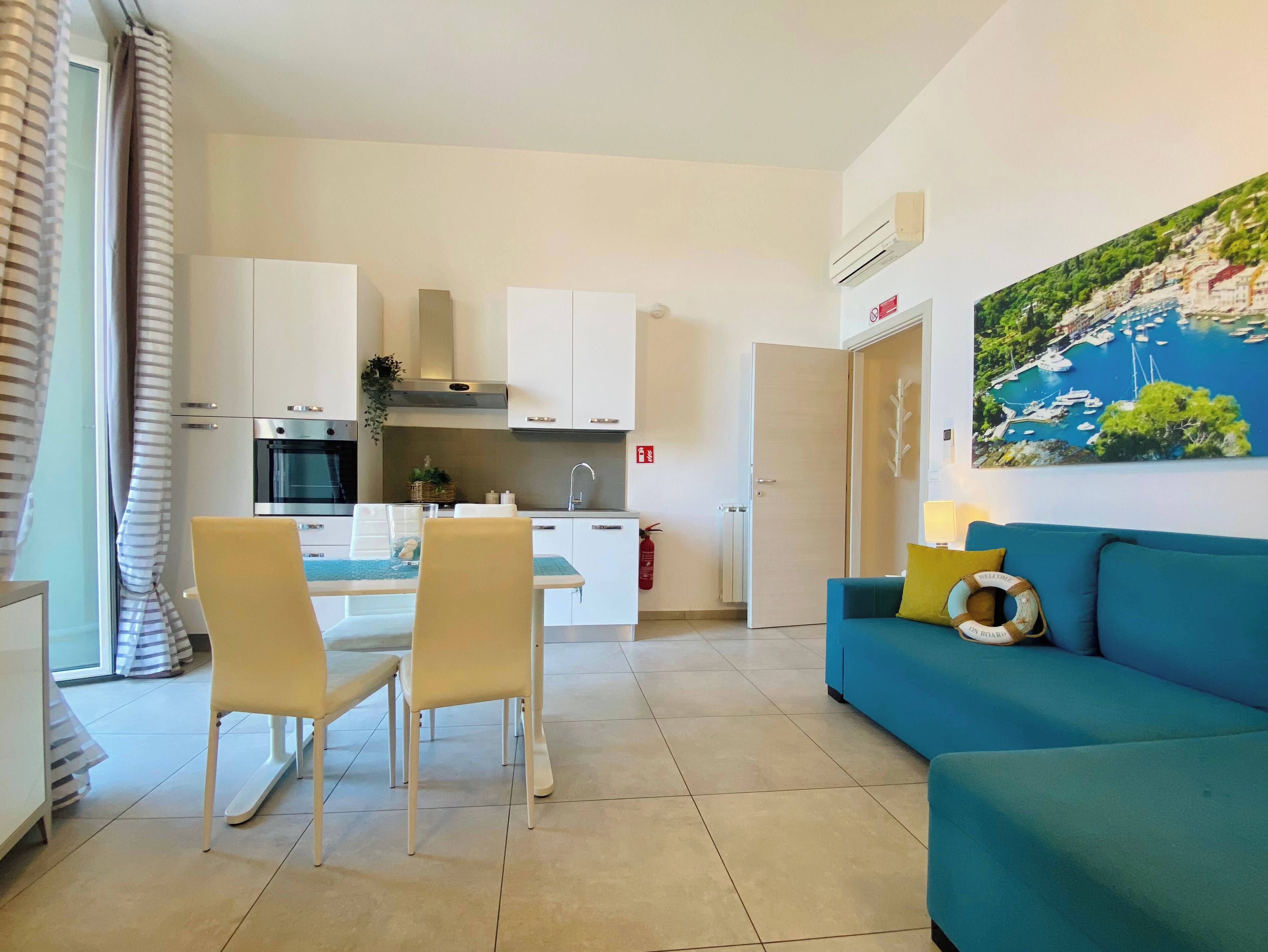 Una cocina moderna con armarios blancos, una zona de comedor con dos sillas y un sofá azul con un cojín decorativo.