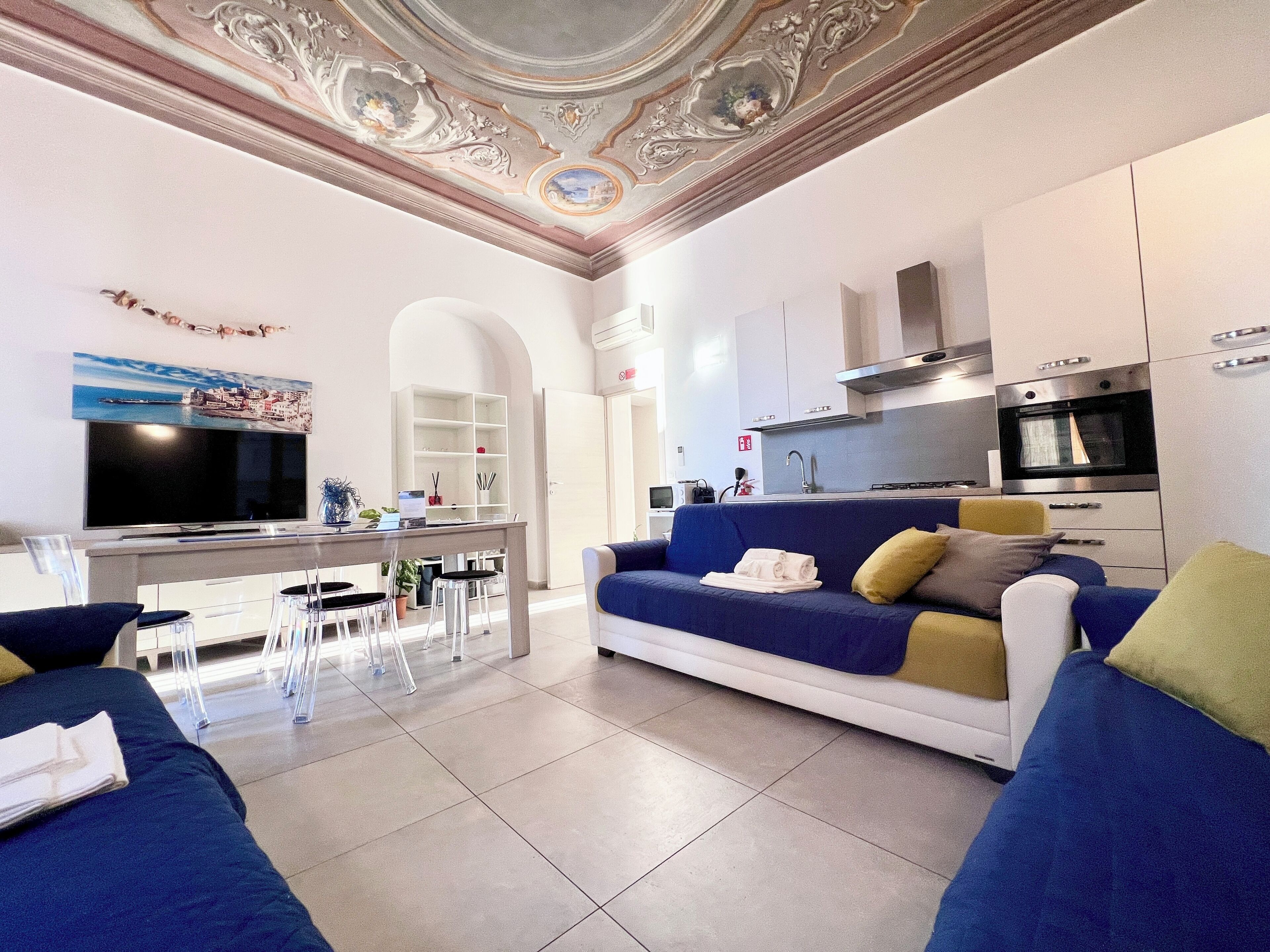 A modern living room with a blue sofa, a television, a kitchen area with a microwave and oven, and a dining area with a table and chairs.