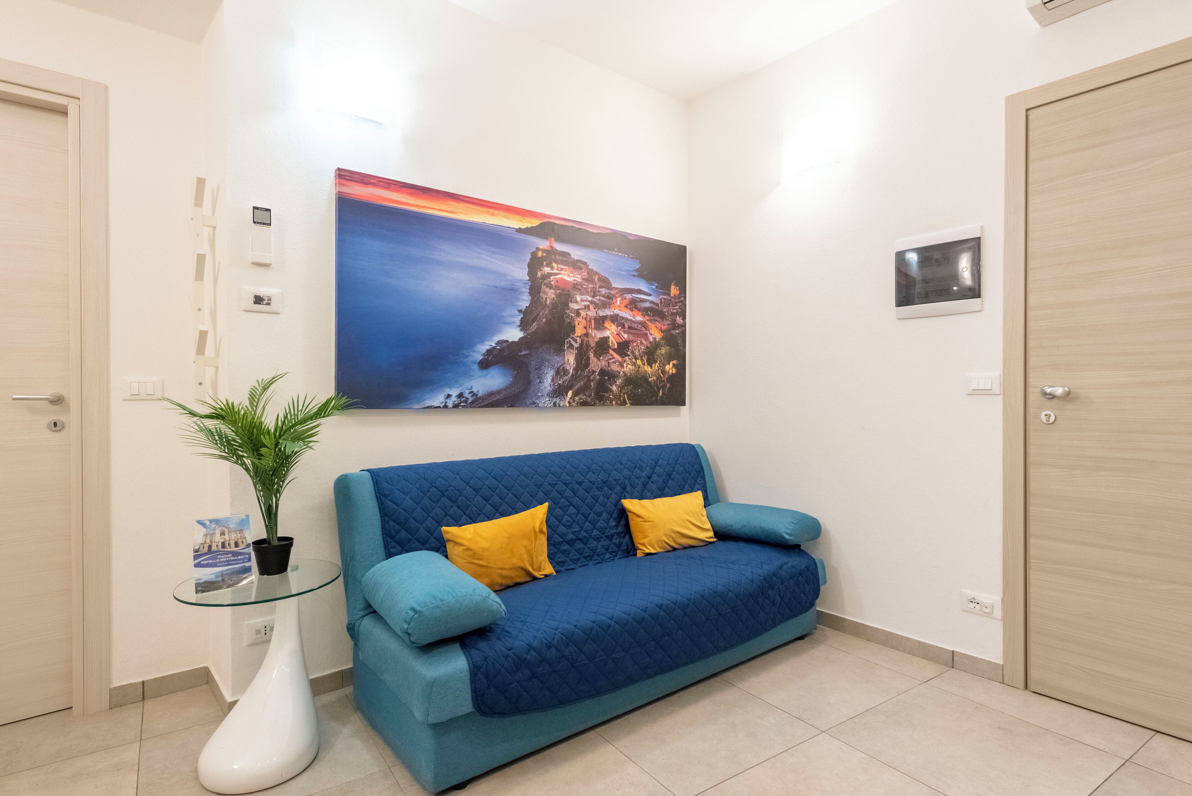 Ein Zimmer mit einem blauen Sofa, einem Glastisch mit einer Pflanze und einem Bild einer Küstenlandschaft.