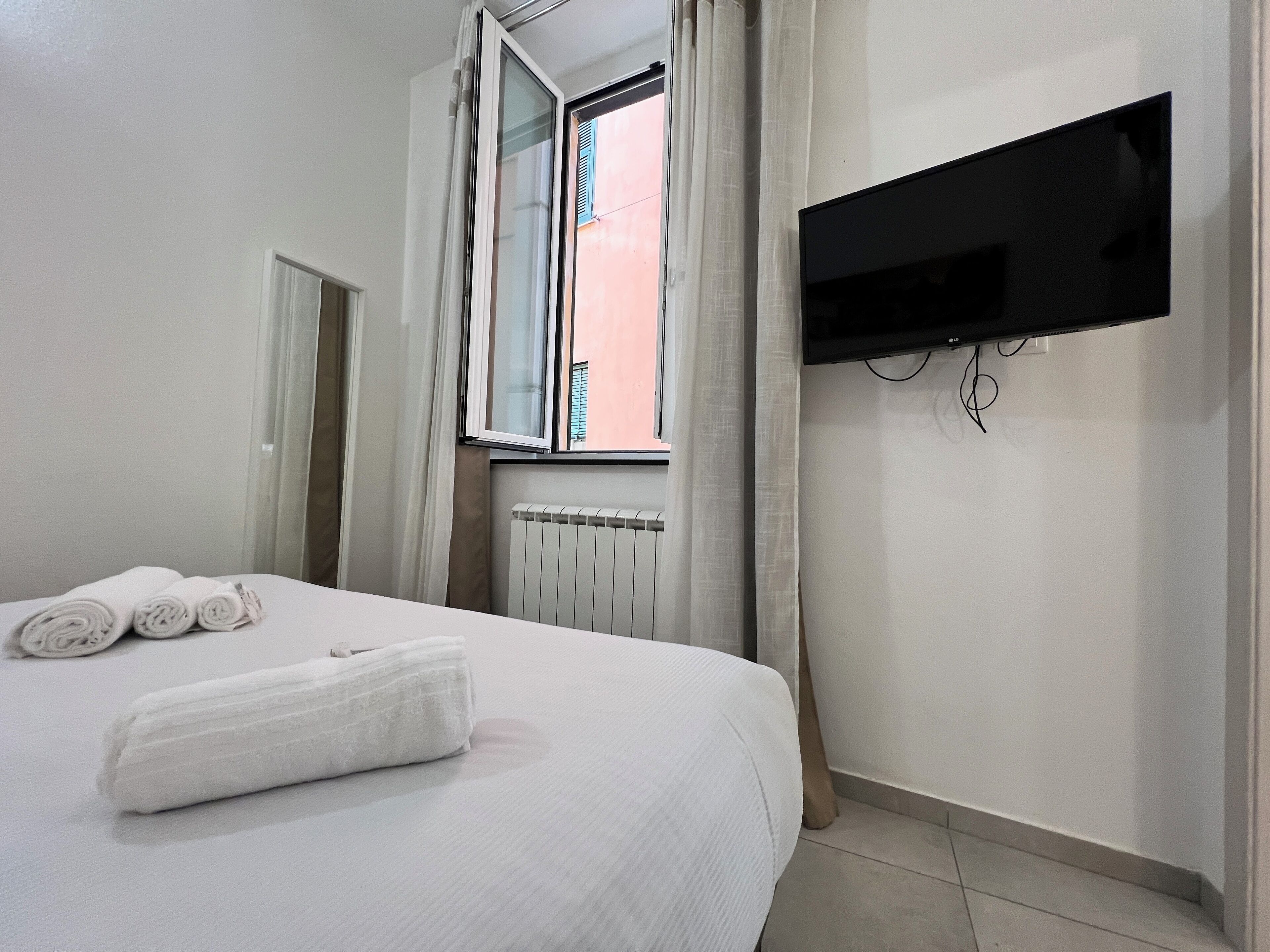 Een hotelkamer met een bed, opgerolde handdoeken, een raam met gordijnen, een radiator en een wandgemonteerde televisie.