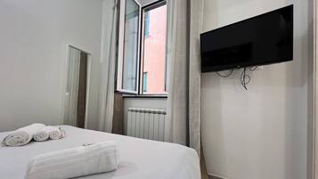 Een hotelkamer met een bed, opgerolde handdoeken, een raam met gordijnen, een radiator en een wandgemonteerde televisie.