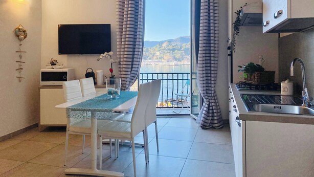 A modern kitchen with a sink, stove, and a dining area with a table and chairs. A TV is mounted on the wall, and there is a view of a body of water and hills in the background. - Casa Rosa al Mare, Rapallo (Rapallo)