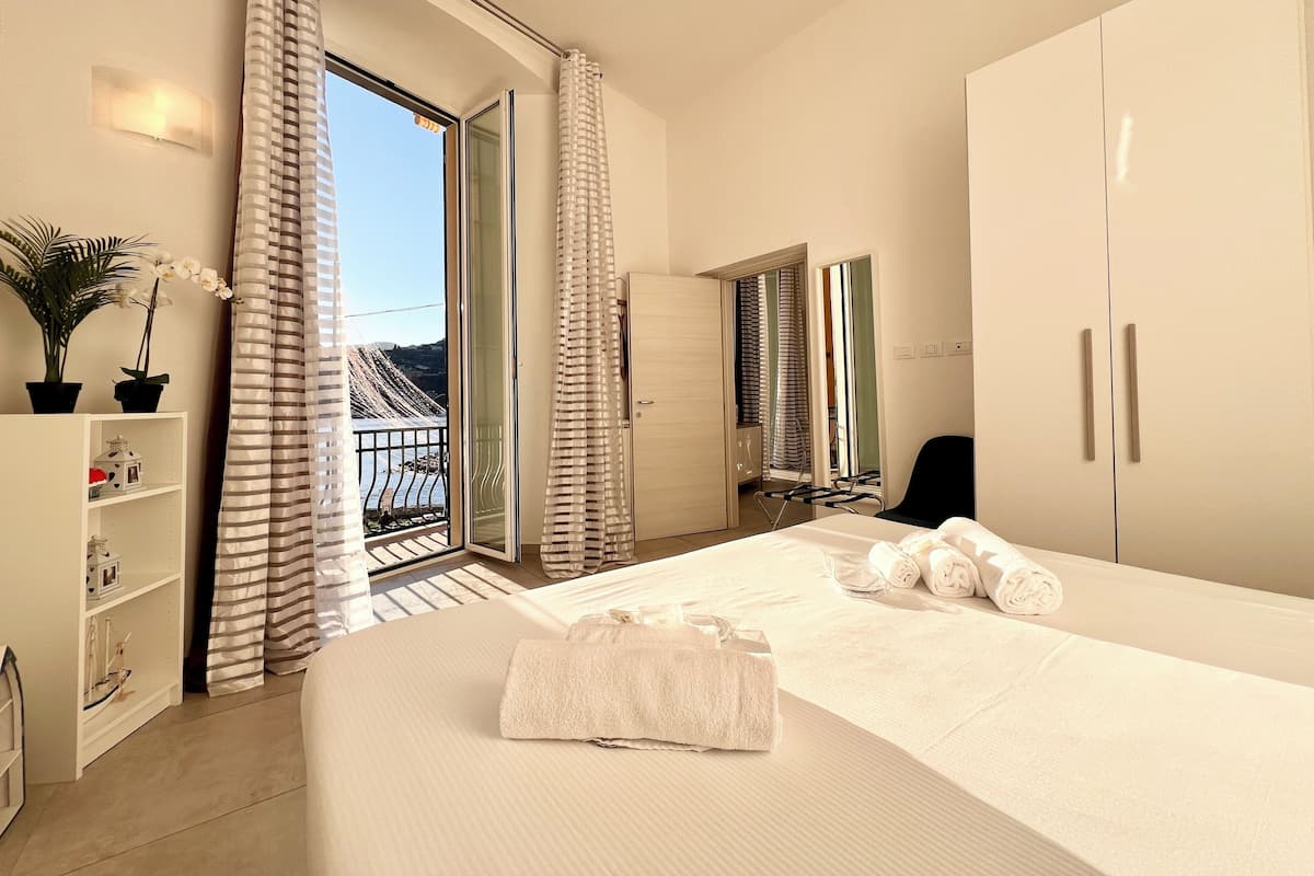 A hotel room with a bed, a wardrobe, a small desk, a chair, a potted plant, and a view of the outdoors.