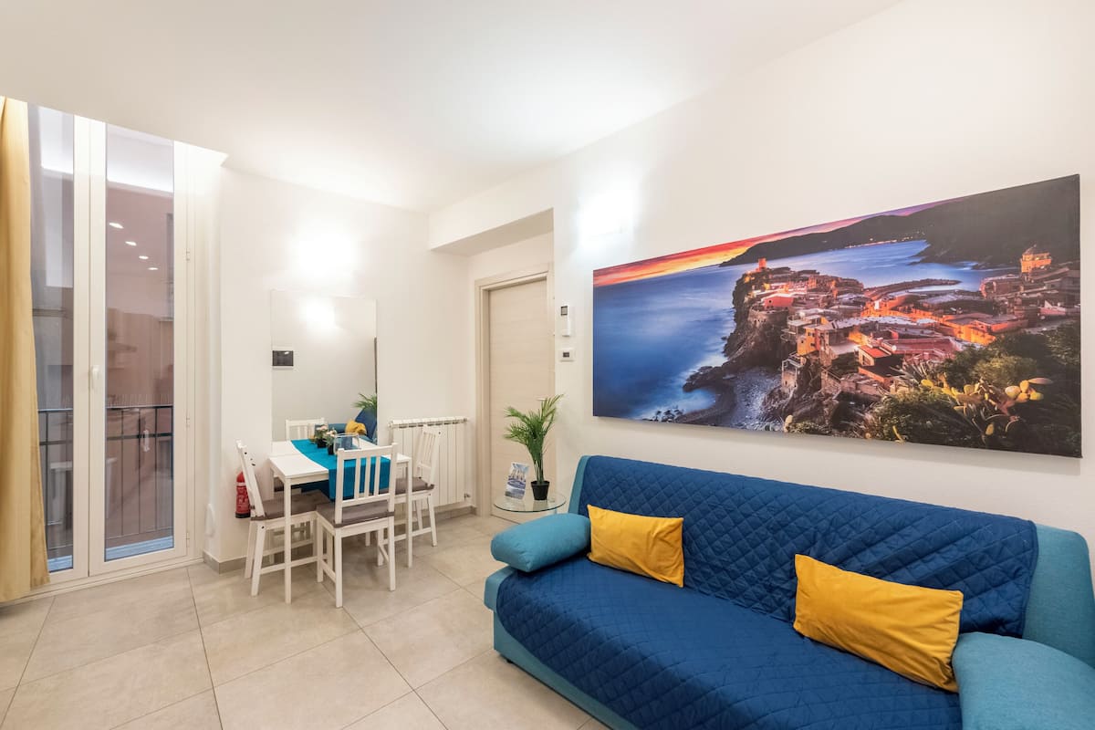 A living room with a blue sofa, a dining table with chairs, and a large painting of a coastal scene.
