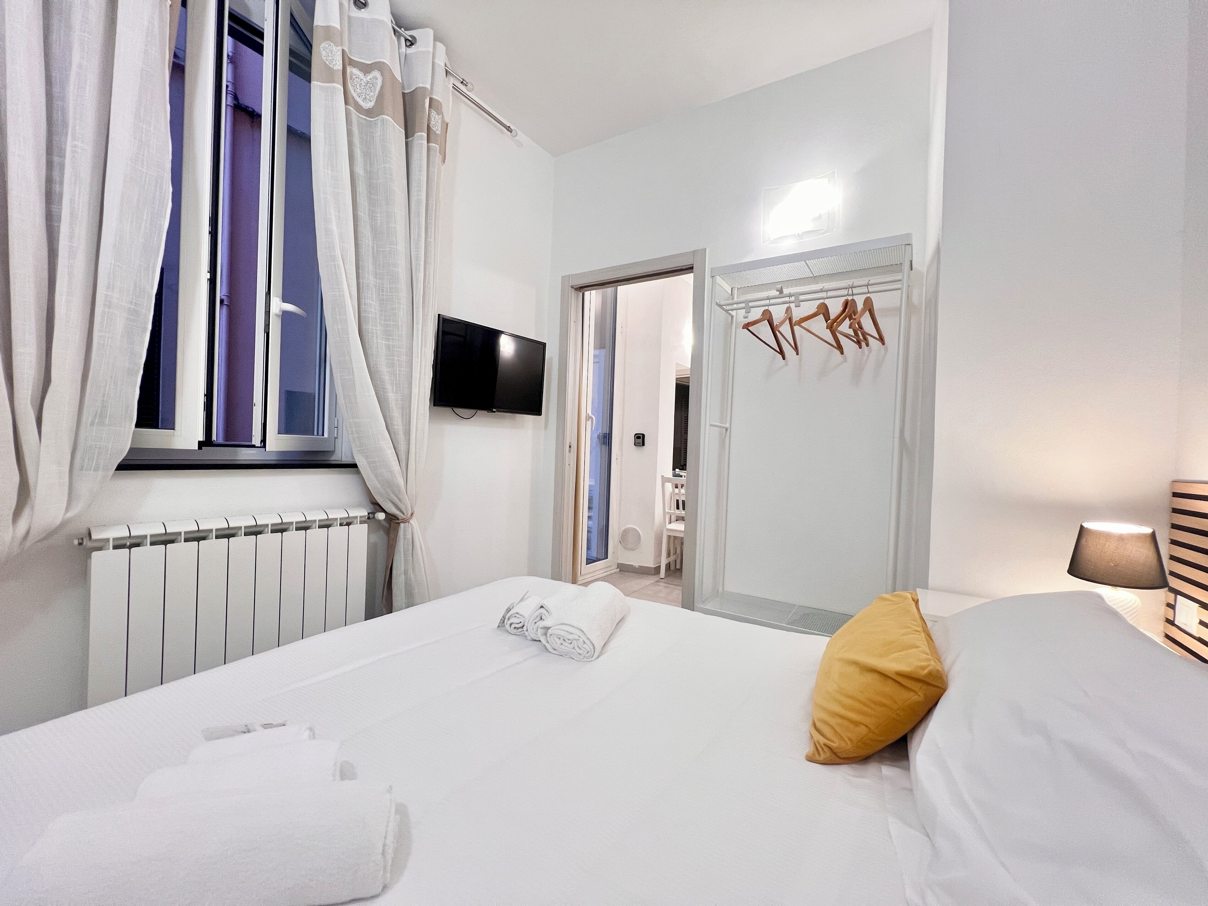Una habitación de hotel con cama, televisión, radiador y ventana con cortinas.