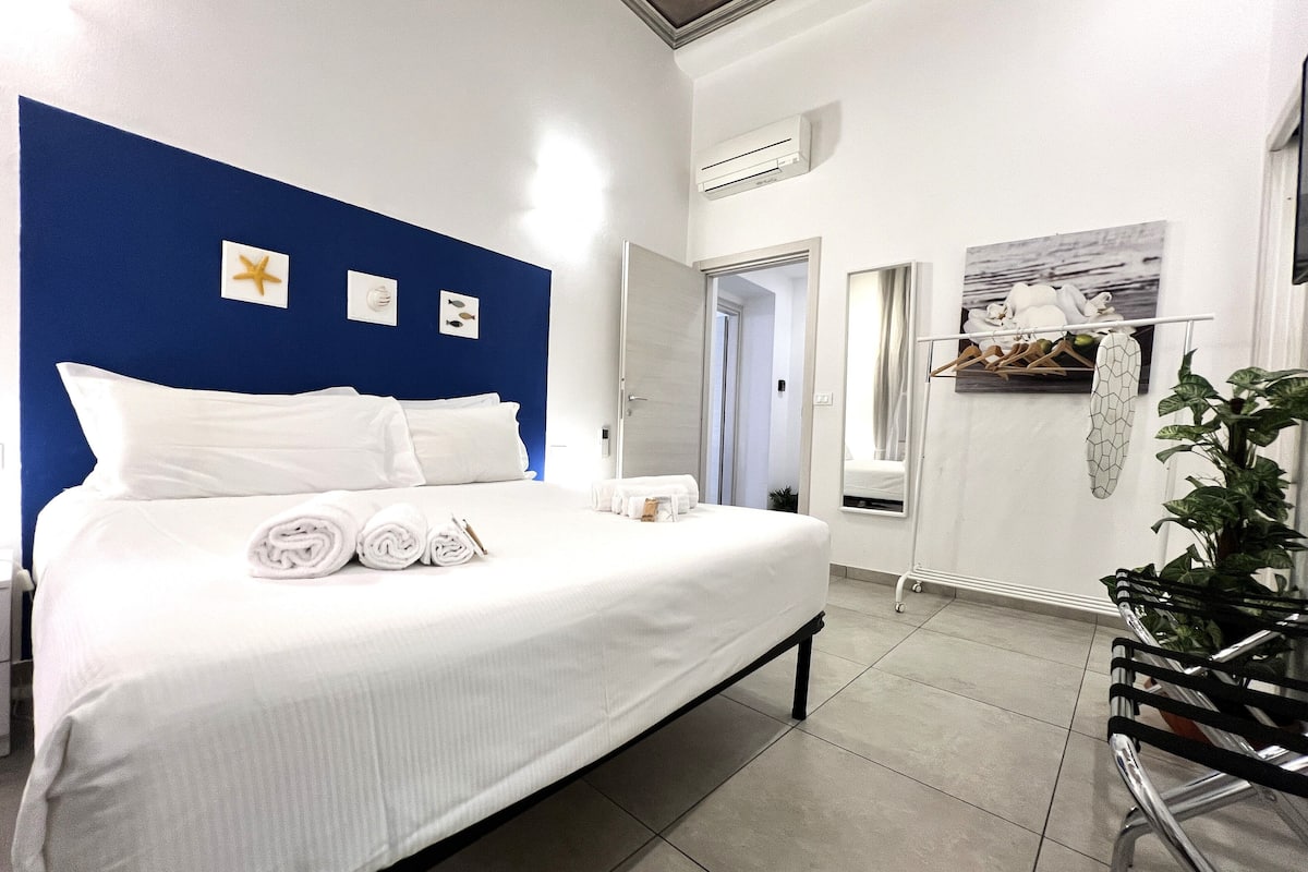 A hotel room with a large bed, white linens, a blue accent wall, a mirror, a plant, and a window.