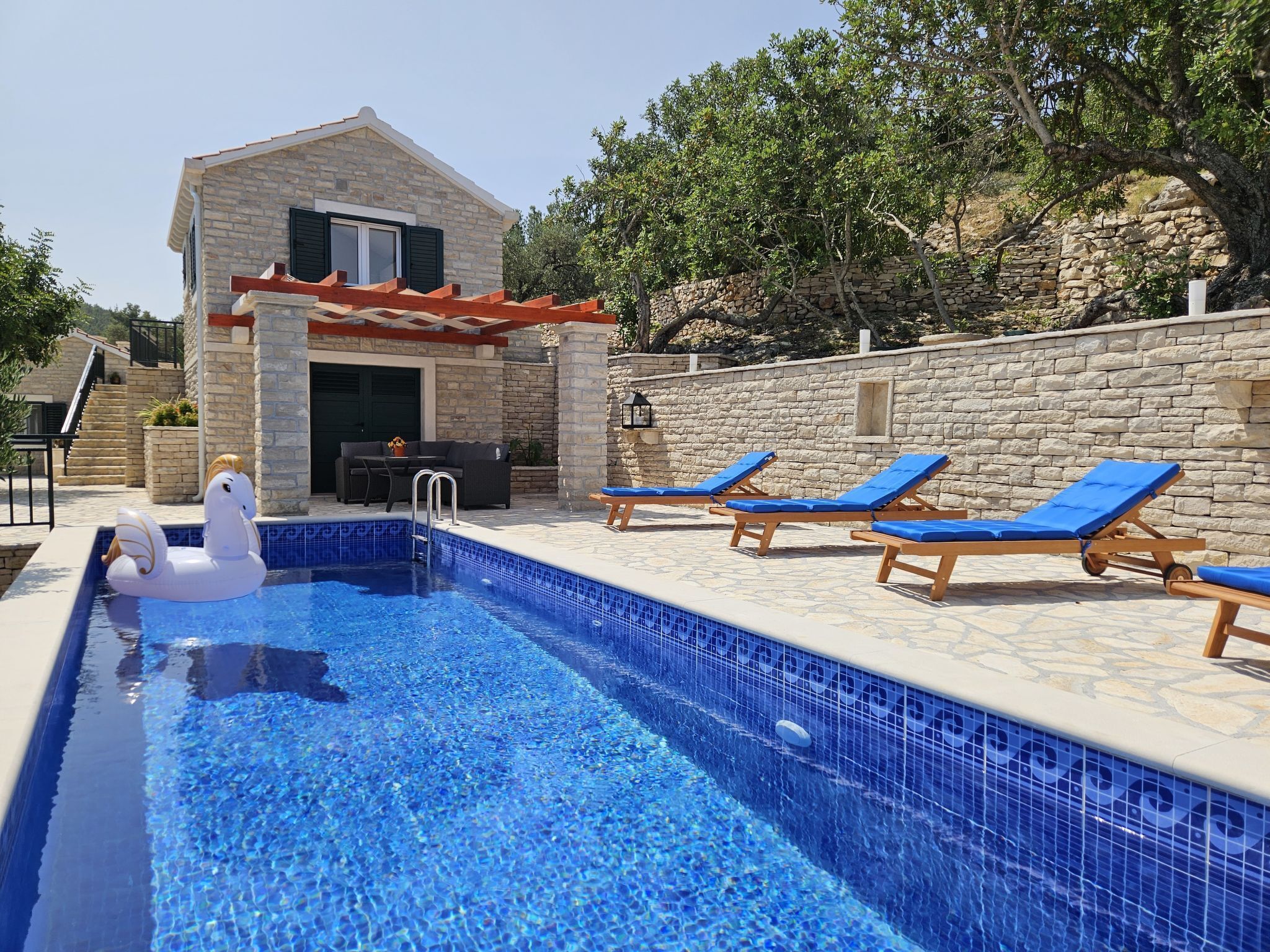 A swimming pool with a swan float, a stone house with a red roof, and wooden lounge chairs.