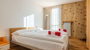 Una habitación de hotel con suelo de madera, una cama con sábanas blancas, cabecero de madera, mesita de noche, lámpara y una ventana con cortinas.