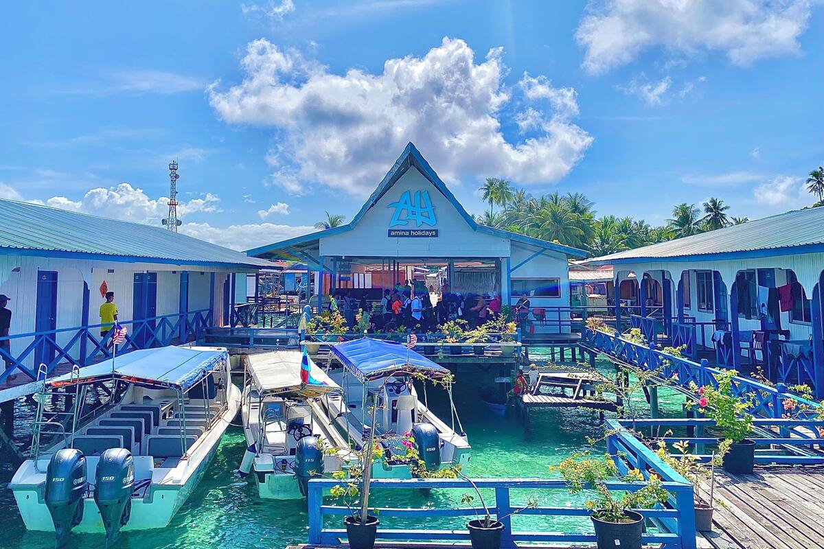 A waterfront hotel with a prominent sign for 'Amina Holidays'. There are boats docked in the water, and a covered walkway with potted plants.