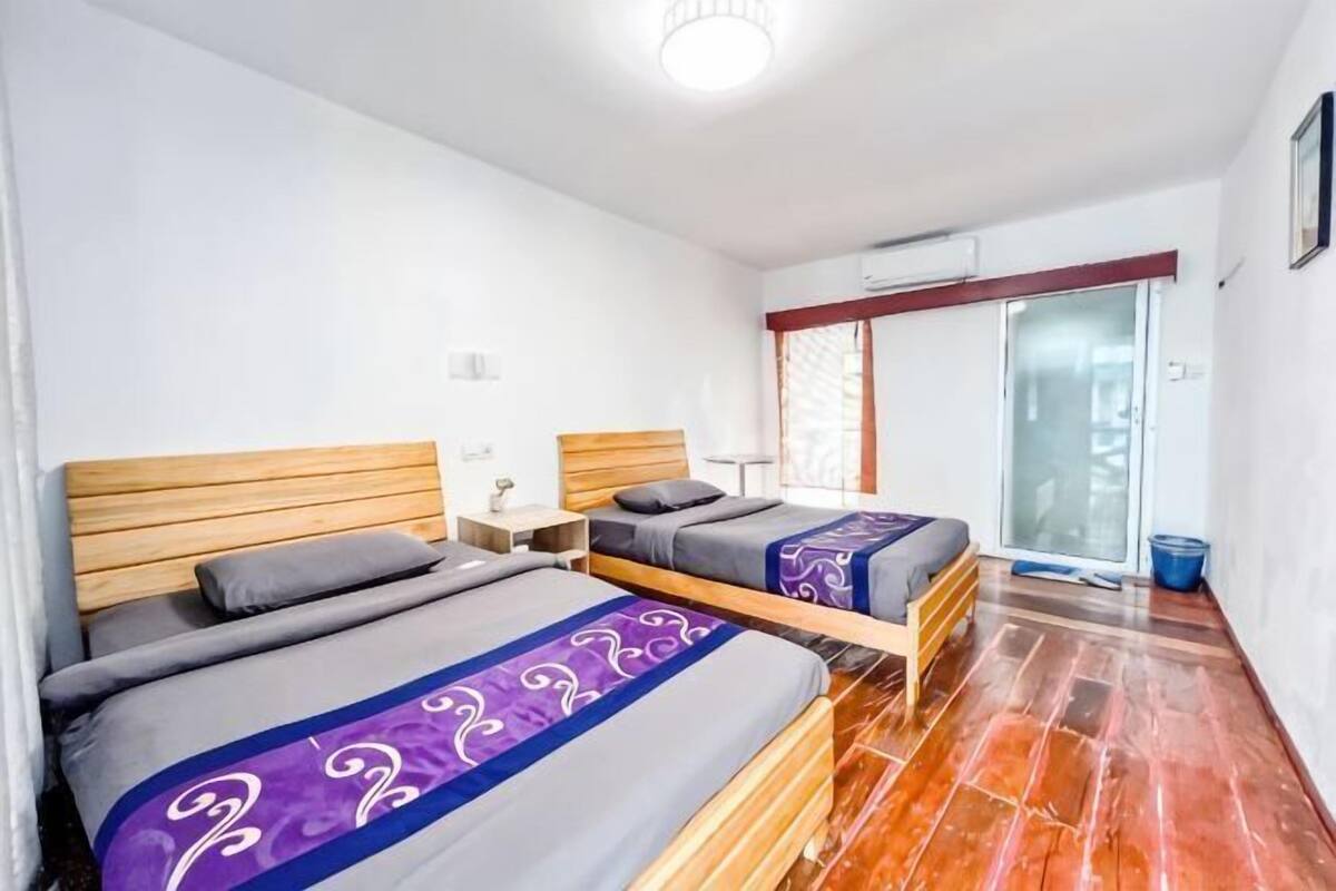 A room with two beds, wooden headboards, a small table, and a sliding glass door leading to a balcony.