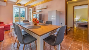 A dining area with a wooden table, four chairs, and a vase with flowers. There is a kitchen area with a refrigerator and cabinets in the background.