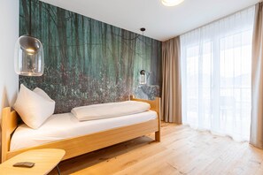 Une chambre moderne avec un lit en bois, une petite table, une lampe fixée au mur et une grande fenêtre ornée de rideaux légers.