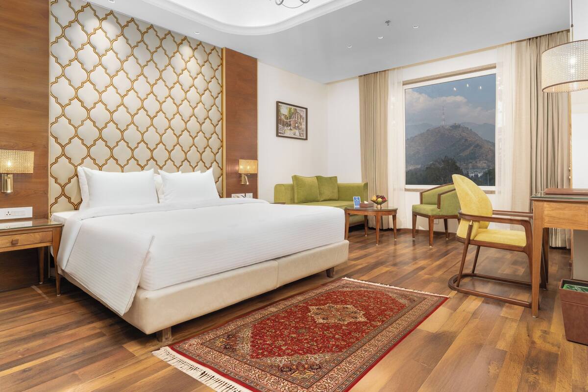 Habitación de hotel con una cama grande, un sofá verde, un escritorio de madera y vistas a las montañas.