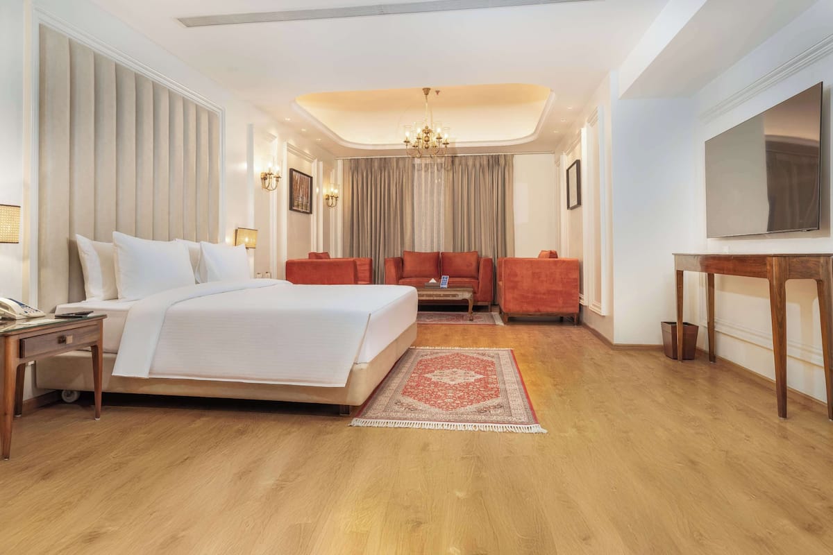 Habitación de hotel con una cama grande, un área de descanso con dos sillones, una mesa de centro de madera, un televisor y una araña.