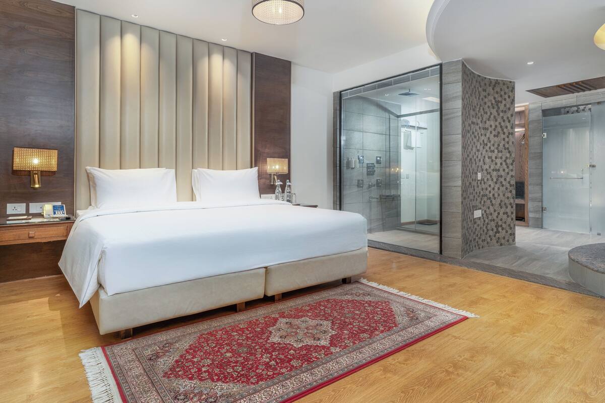 Una habitación de hotel moderna con una cama grande, lámparas de noche, un box de ducha con vidrio y suelo de madera.