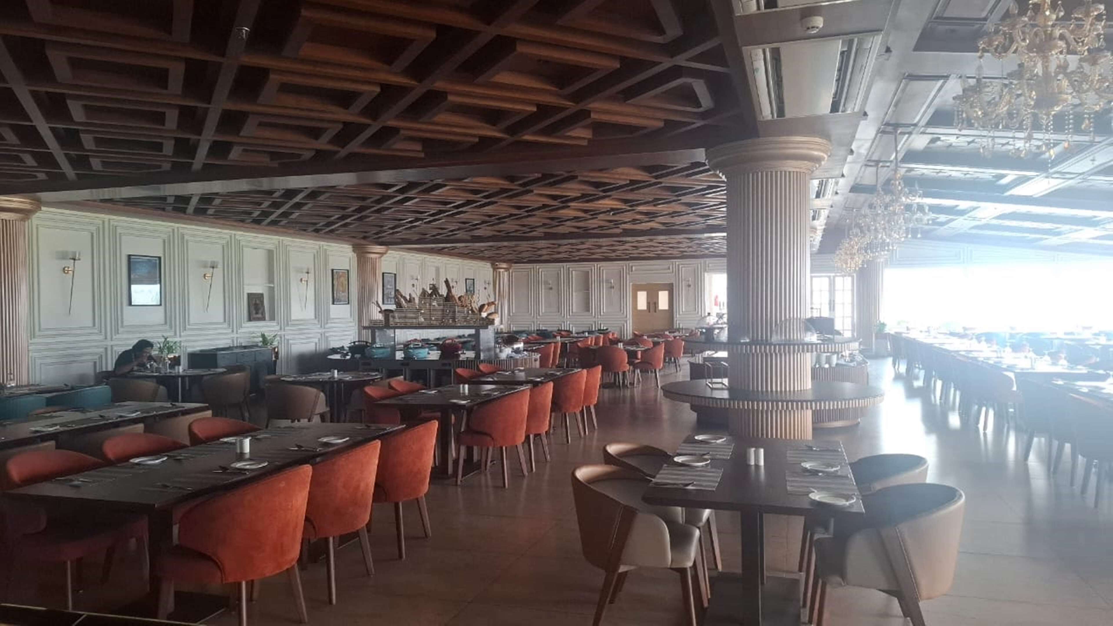 A spacious restaurant with high ceilings, wooden beams, and a central column. Tables are arranged in rows, and there are chairs around each table.