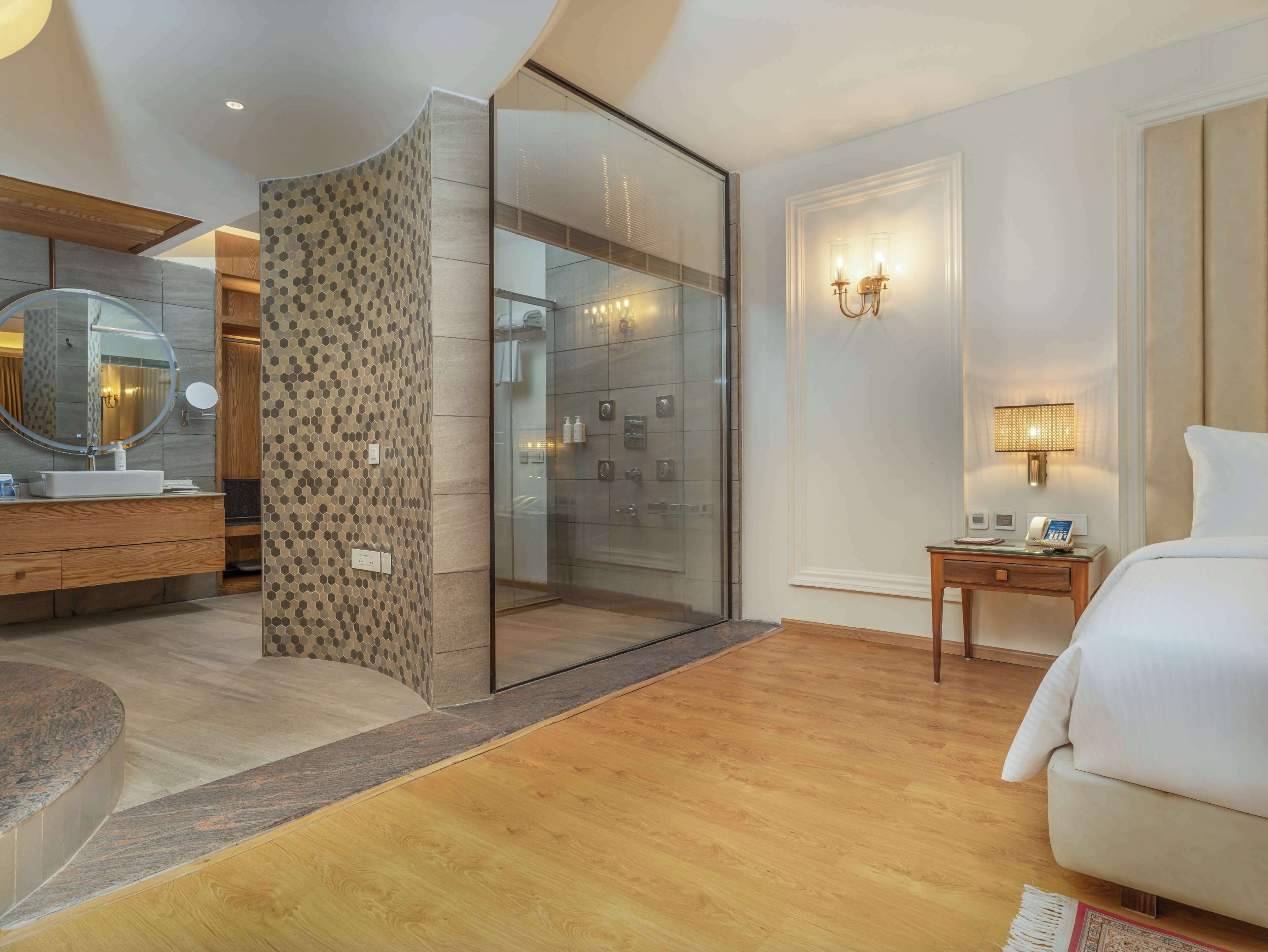 Una habitación de hotel moderna con cabina de ducha de vidrio, tocador de madera, un espejo grande, una mesita de noche con lámpara y una cama.