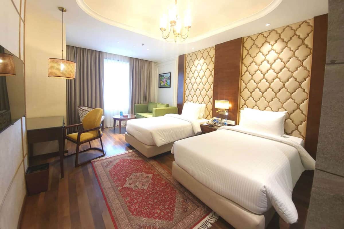 Habitación de hotel con dos camas, zona de estar con una silla y una mesa, un ventanal grande con cortinas y un panel decorativo en la pared.
