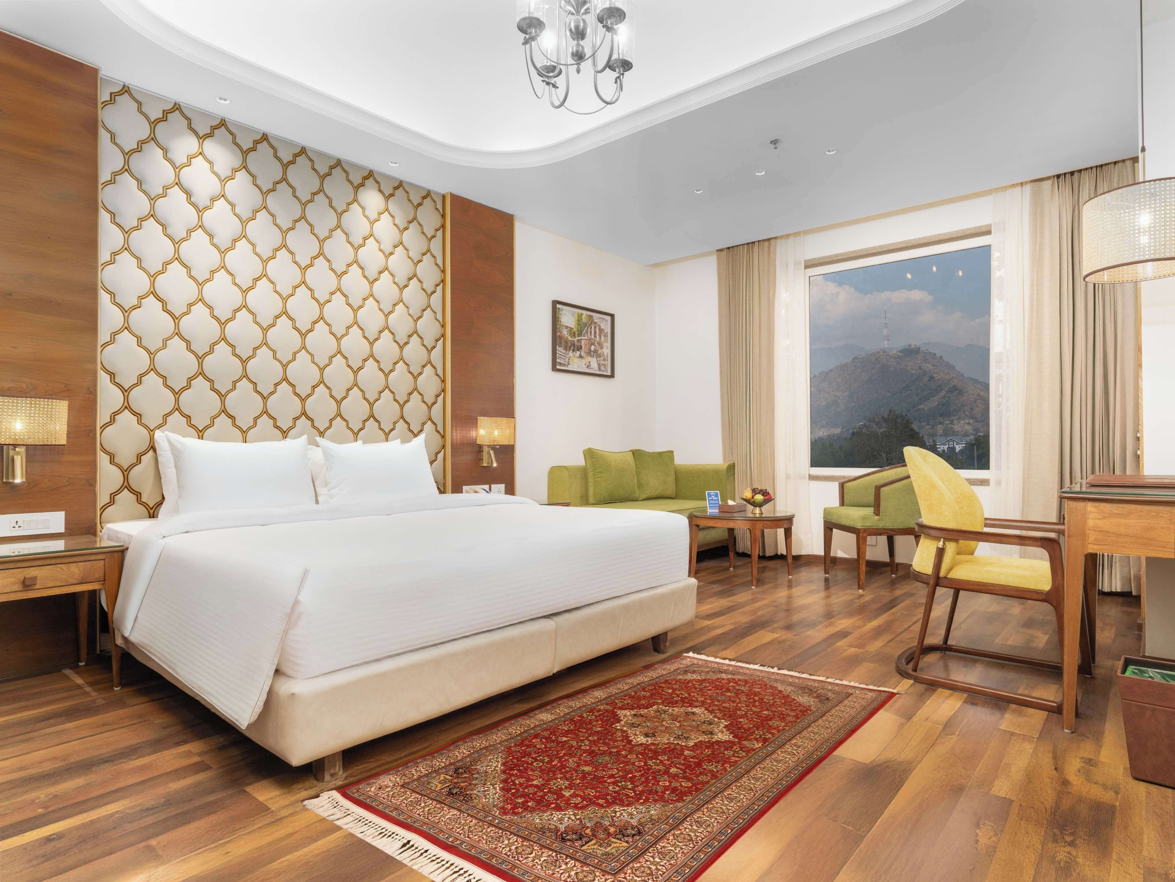 A hotel room with a large bed, a green sofa, a small table with a fruit bowl, a desk, and a view of mountains through the window.