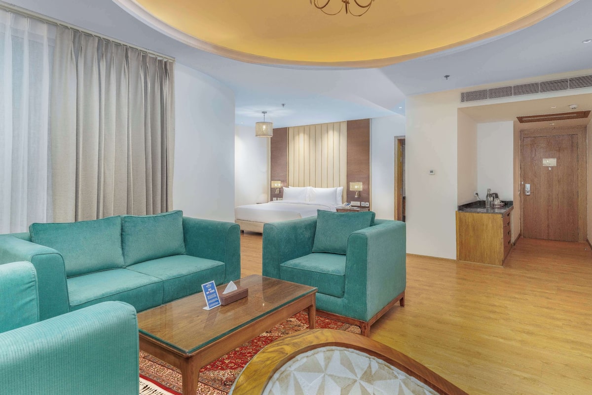 Una habitación de hotel con una cama grande, dos sofás color turquesa, una mesa de centro de madera y un candelabro.