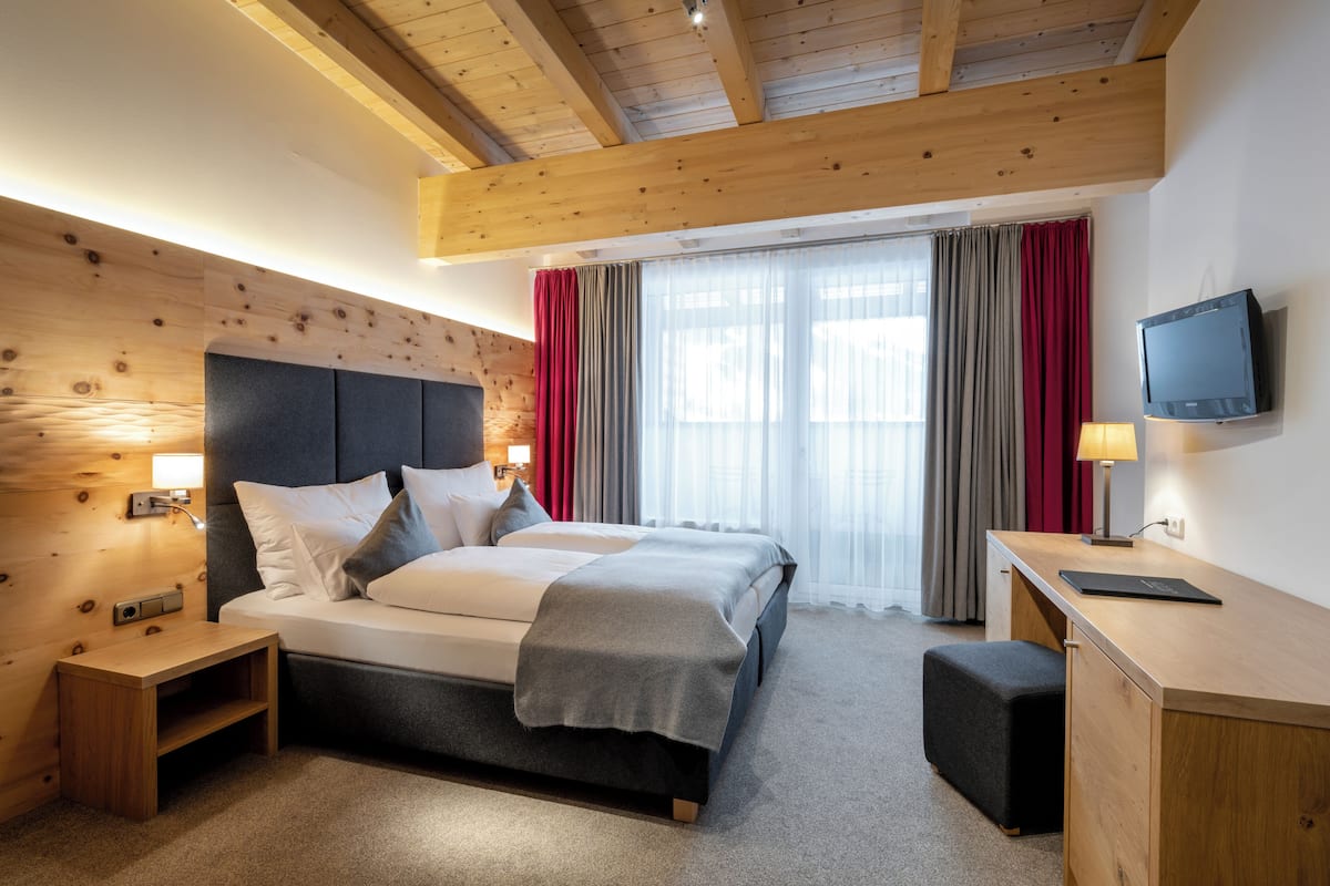 A modern hotel room with a wooden ceiling, a large bed with a grey headboard, a desk with a lamp, and a TV mounted on the wall.