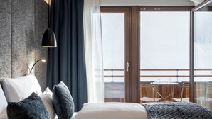 Habitación de hotel moderna con una cama grande, cortinas oscuras, vistas al balcón y una mesa de madera con sillas.