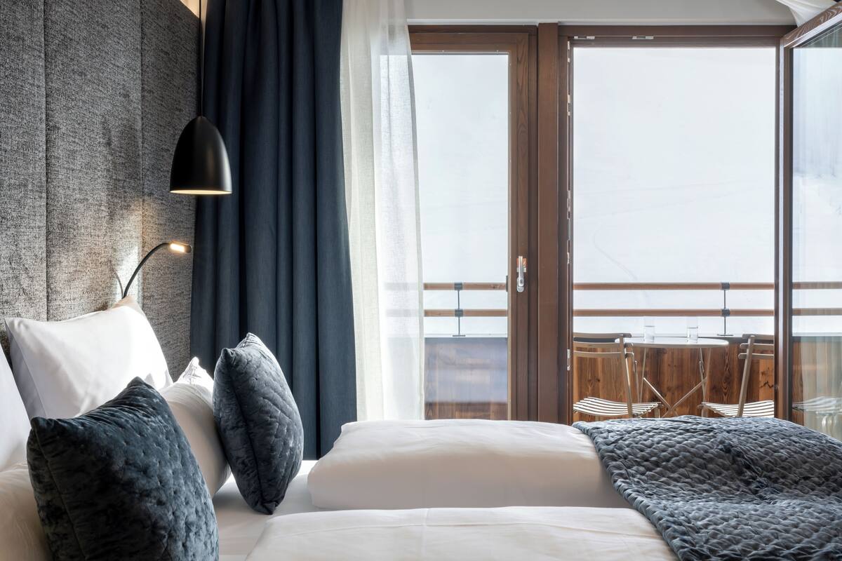 A modern hotel room with a large bed, dark curtains, a view of the balcony, and a wooden table with chairs.