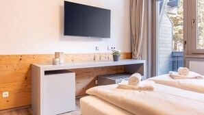 A hotel room with a flat-screen TV mounted on the wall, a wooden accent wall, a small desk with a plant, a mini-fridge, and two beds with towels.