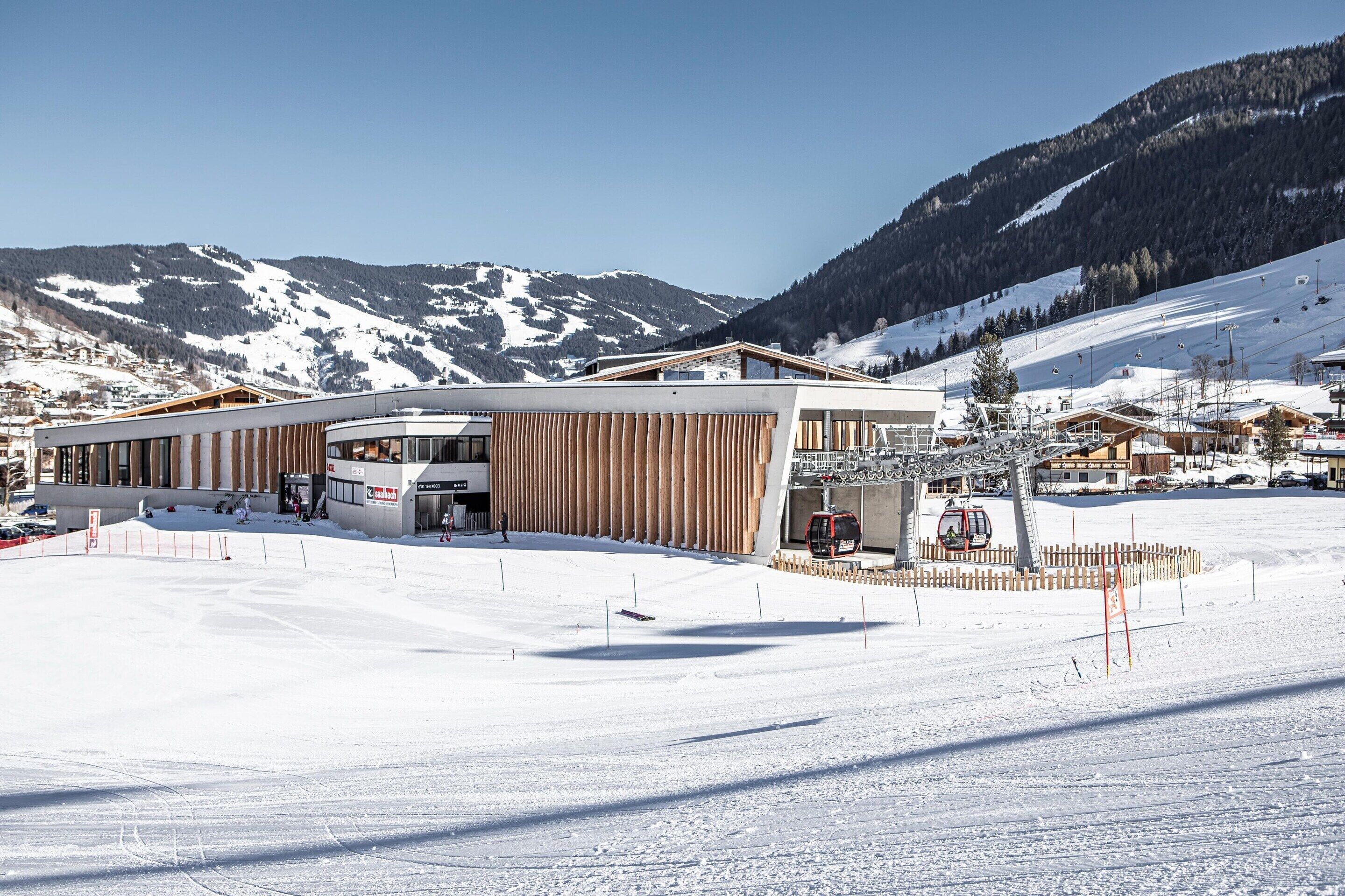 A modern ski resort building with a curved facade, ski lift, and snow-covered mountains in the background.