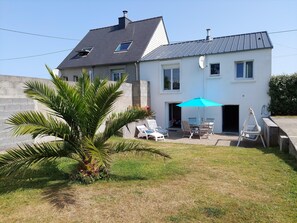 Une maison à deux étages avec un patio comprenant une table et des chaises, un parasol bleu et un palmier.