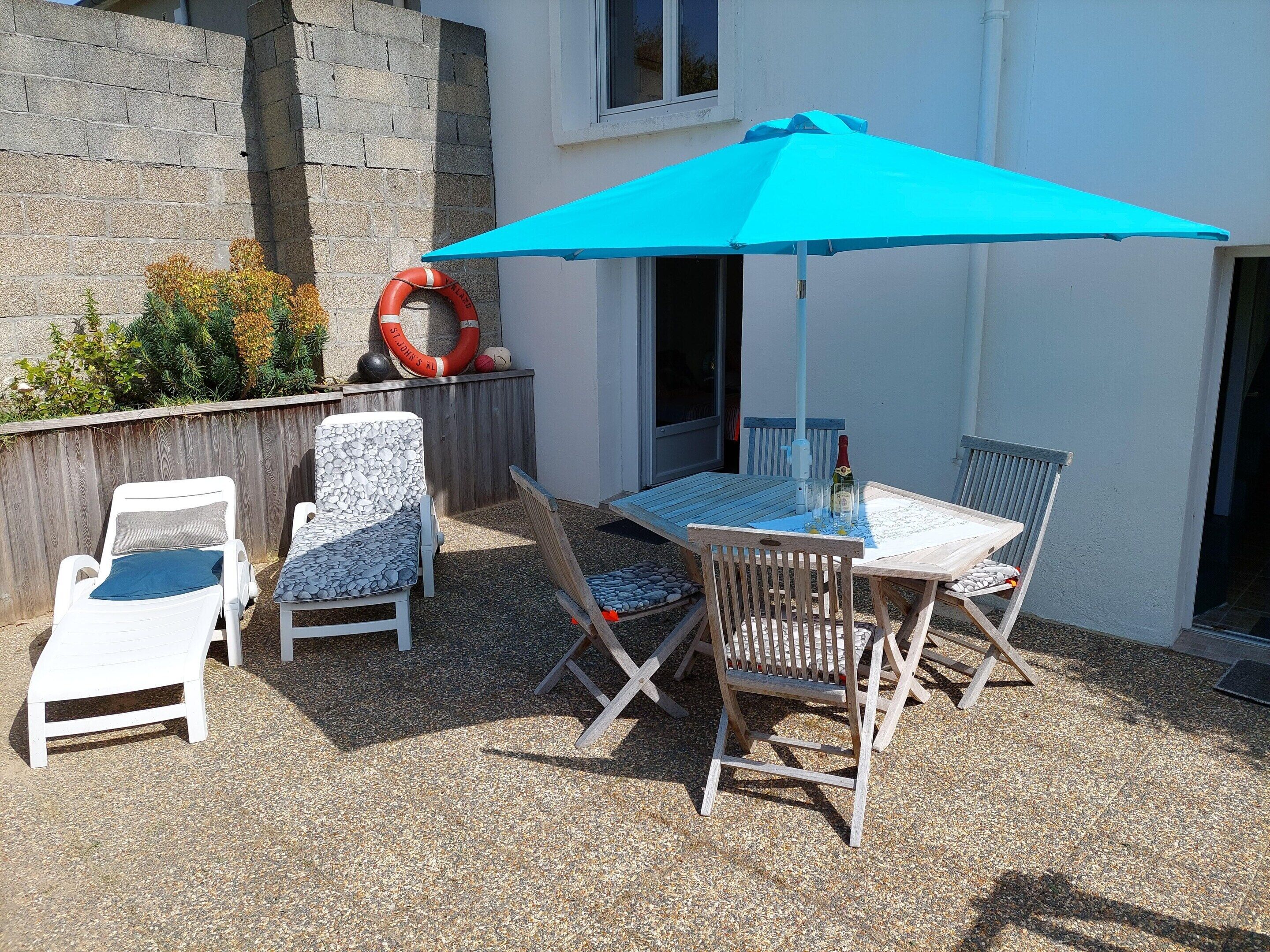 Patio esterno con tavolo e quattro sedie, un ombrello blu e un salvagente appoggiato a una parete.