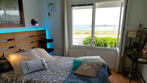 Una camera da letto con un letto rivestito di tessuto effetto pietra, una testiera in legno, una finestra vista spiaggia e un comodino con una lampada.