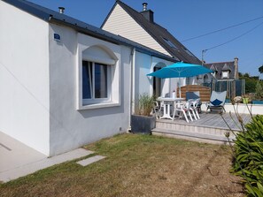Une maison blanche avec un petit patio comprenant une table et des chaises, un parasol bleu et une terrasse en bois.