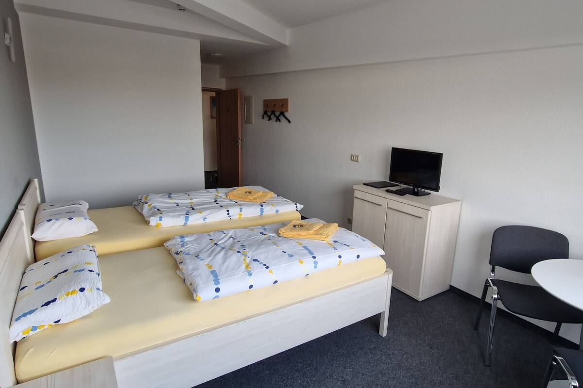 A single bed with a white and blue polka dot duvet, a small desk with a computer, a chair, and a round table with a glass.