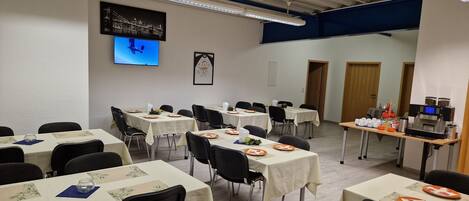 Un salón de conferencias con varias mesas dispuestas para comer, un televisor en la pared y una estación de café.