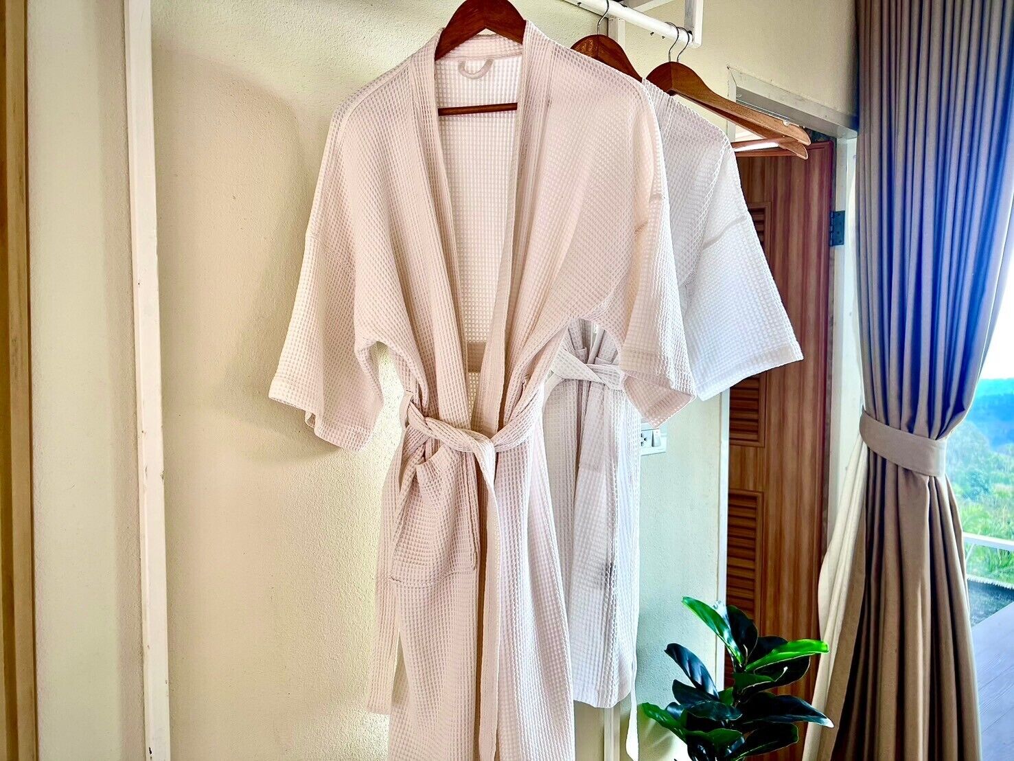 A white bathrobe hanging on a wooden hanger in a room with a view of greenery through a window.