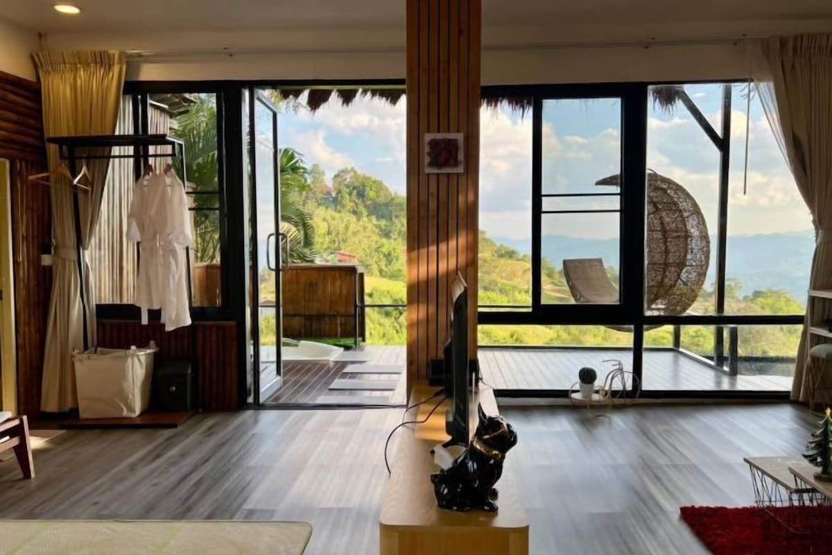 Une pièce spacieuse dotée d’une grande porte-fenêtre coulissante donnant sur un balcon avec vue sur la verdure et les montagnes. On y trouve un plancher en bois, une petite table et une chaise.