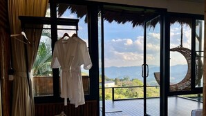 A room with a large sliding glass door leading to a balcony with a view of mountains and greenery. There is a white robe hanging on a rack and a white bag on the floor. - Bella Lunar Villa (Mae Fa Luang)