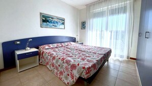 A bedroom with a bed, a nightstand, a desk, a chair, a window with curtains, and a painting on the wall. - Relaxing Coastal Stay in Lido Altanea (Caorle (VE))