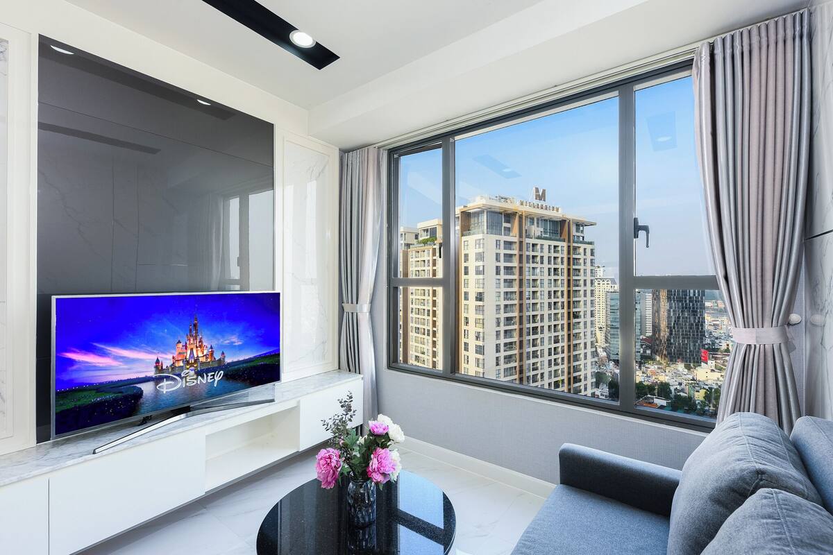 A modern living room with a large TV, a sofa, a small table with flowers, and a large window with a city view.