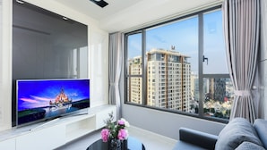 A modern living room with a large TV, a sofa, a small table with flowers, and a large window with a city view. - Amazing CBD Rivergate Apartment & Pool (Ho Chi Minh City)