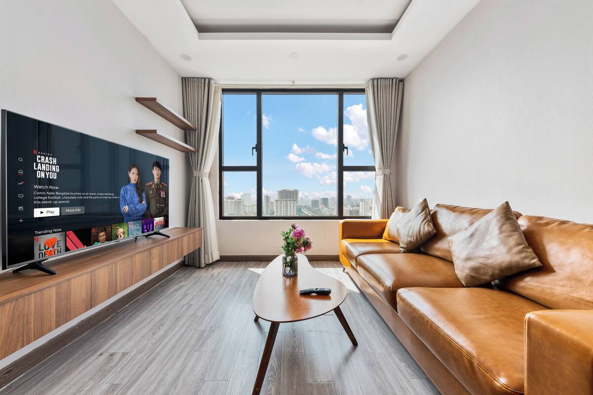 A modern living room with a large TV on a wooden stand, a brown leather sofa, a coffee table with a vase, and a city view through large windows.