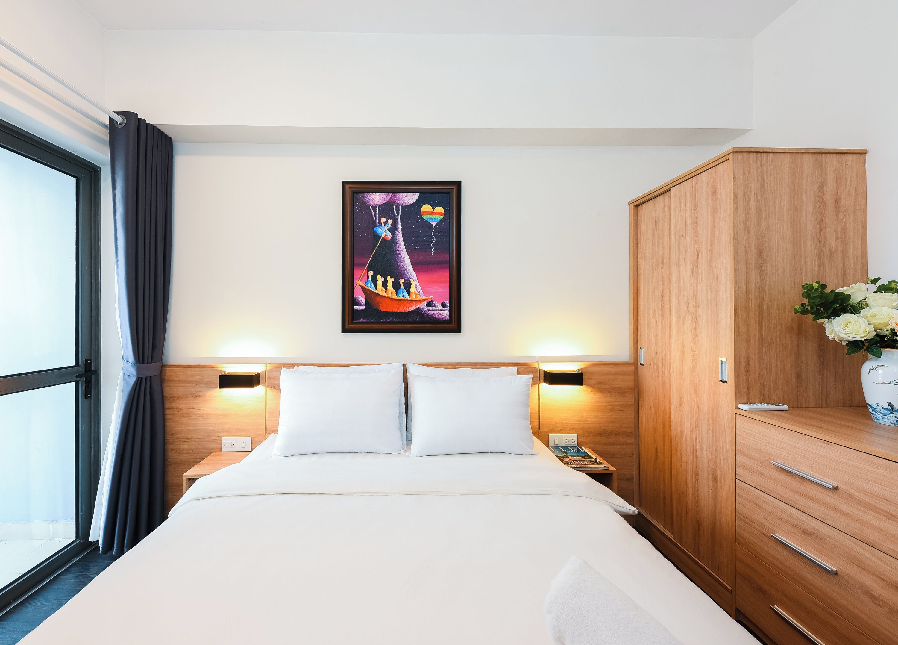 A hotel room with a bed, a wooden wardrobe, a nightstand with a lamp, and a painting on the wall.