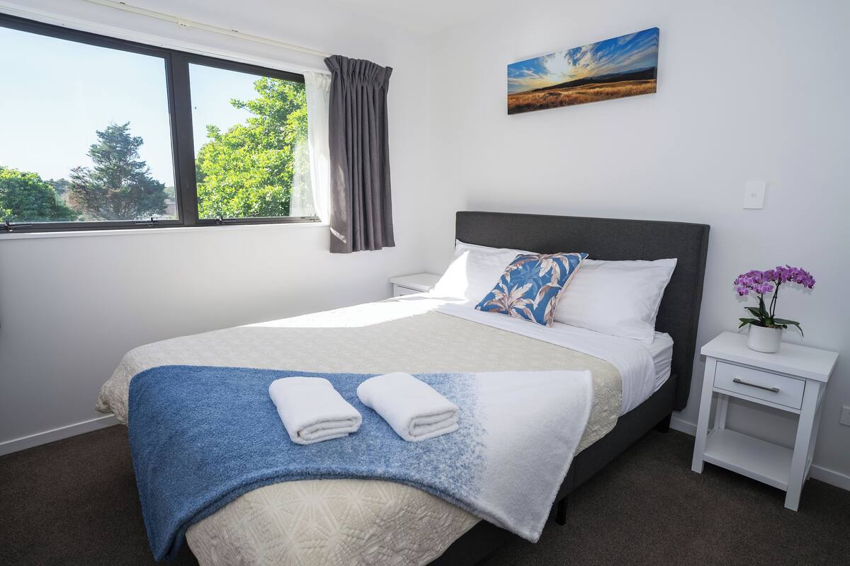 A neatly made bed with a blue and white blanket, a white pillow, and a decorative pillow. There is a small white nightstand with a potted plant beside the bed. A window with curtains is visible, and a painting hangs on the wall.