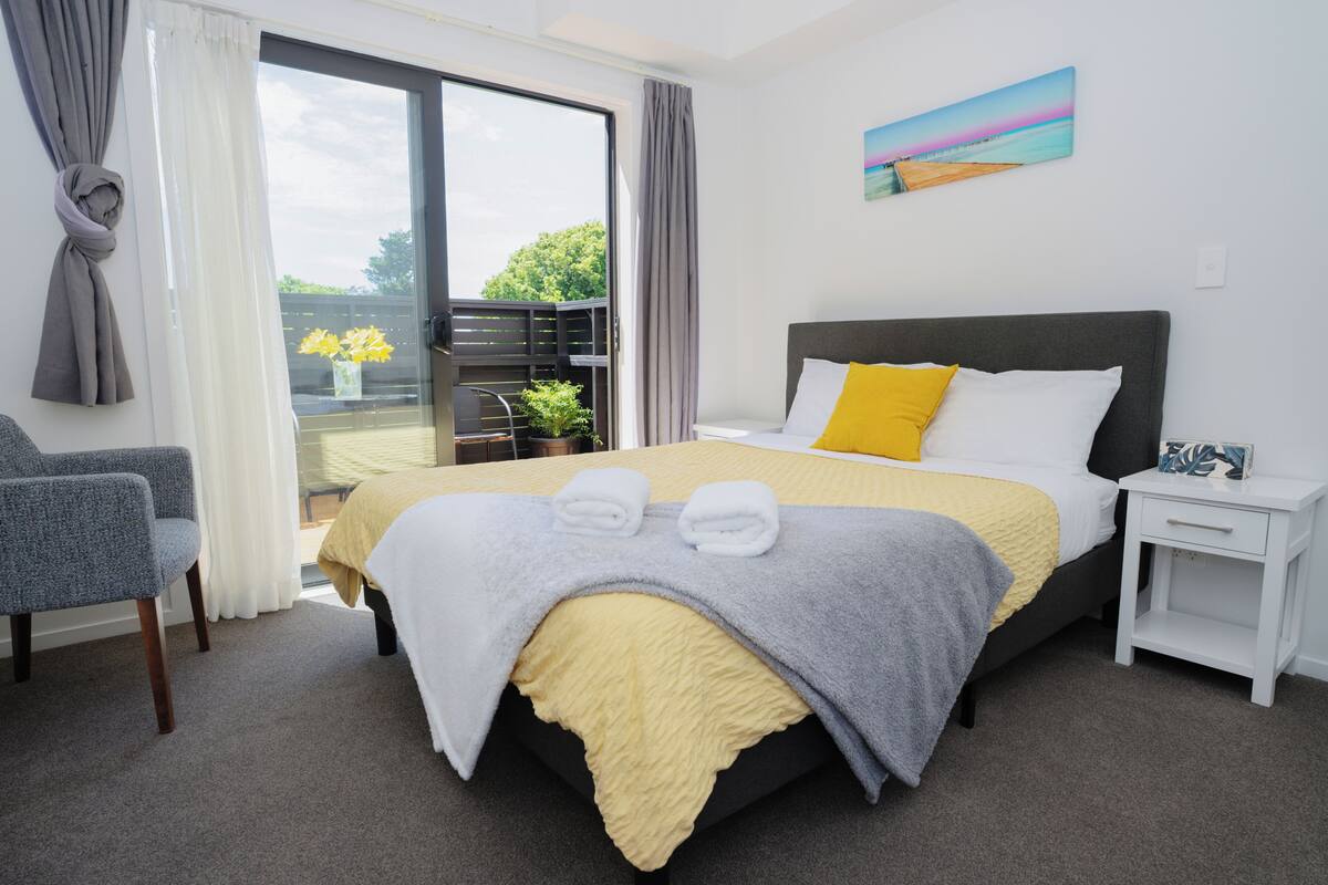 A hotel room with a bed, a chair, a nightstand, and a view of the outdoors.