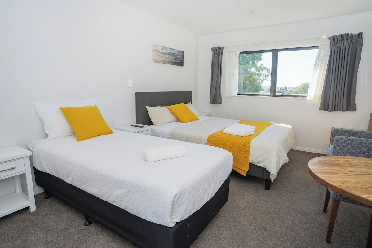 A hotel room with two beds, a small table, a nightstand, and a window with curtains.