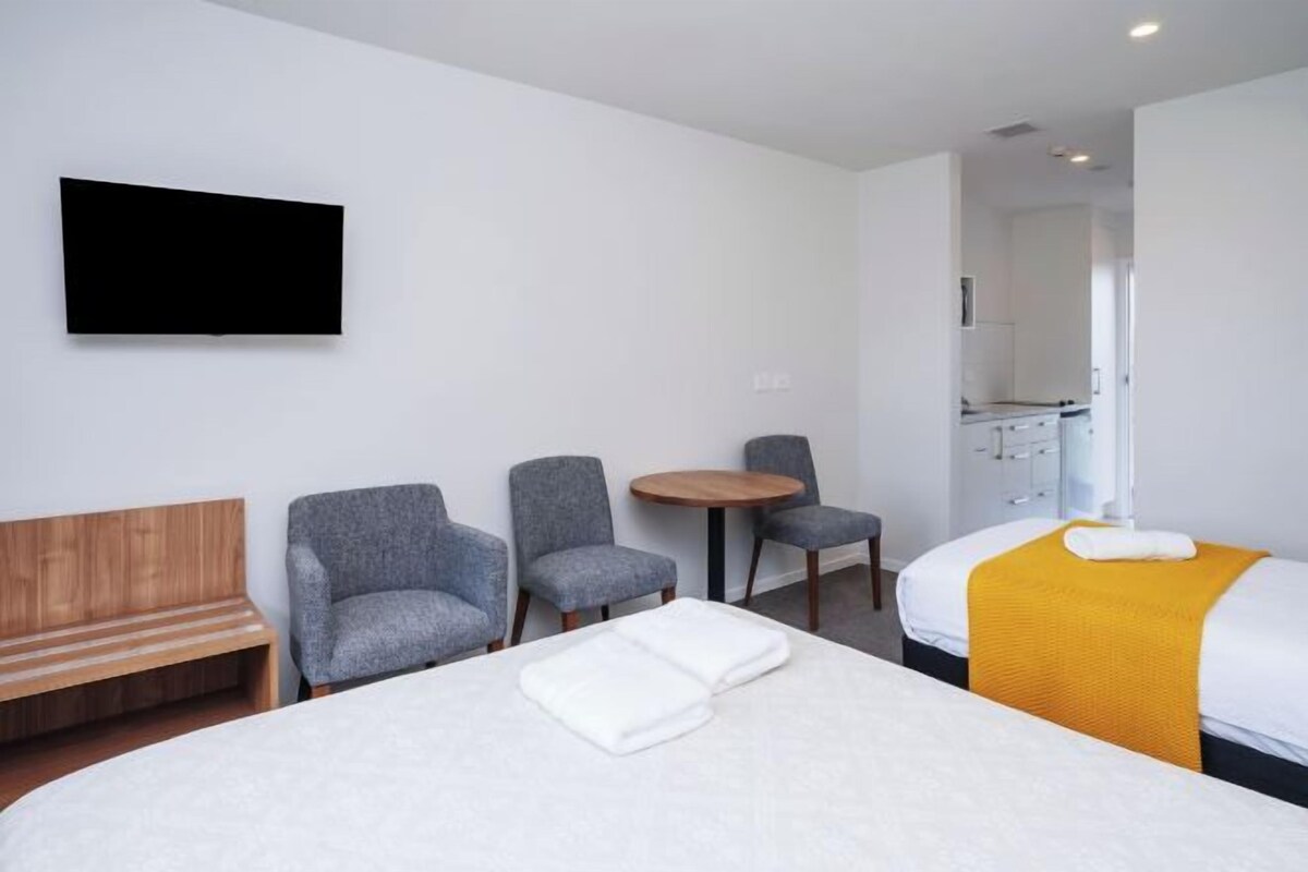 A hotel room with a bed, two armchairs, a small round table, a wooden bench, a TV mounted on the wall, and a bathroom visible through an open door.