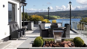 Une terrasse extérieure avec vue sur un lac et des collines, aménagée avec des chaises et une table, et dotée d’un foyer.