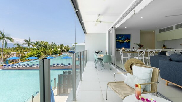 A balcony with a pool, dining area, and living space with a view of palm trees and mountains.